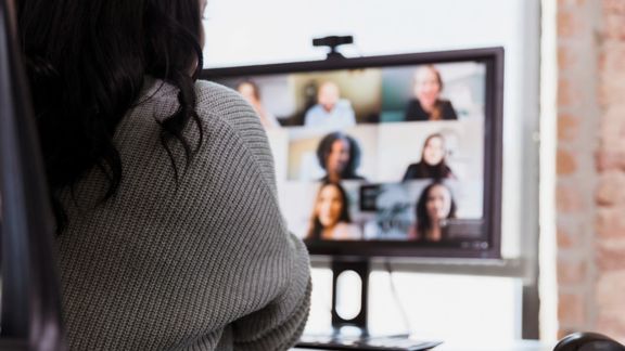 Rear view of a businesswoman on a video conference with a group of colleagues. Focus is on the woman, the computer monitor is blurred.